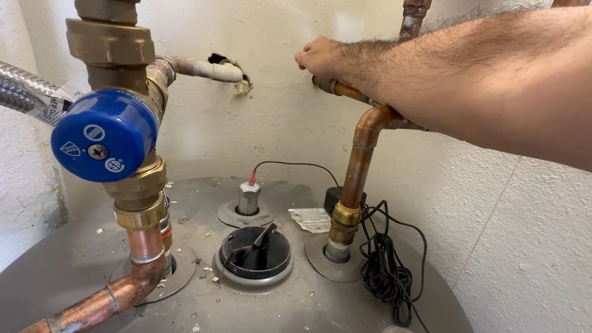 Arm reaching across the top of a gray water heater with a blue valve, copper elbows and a relief fitting