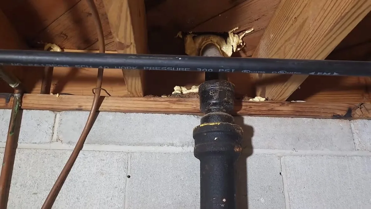 Black cast iron drain stack running down a block wall beneath wooden joists, with a black supply line and copper tube beside it