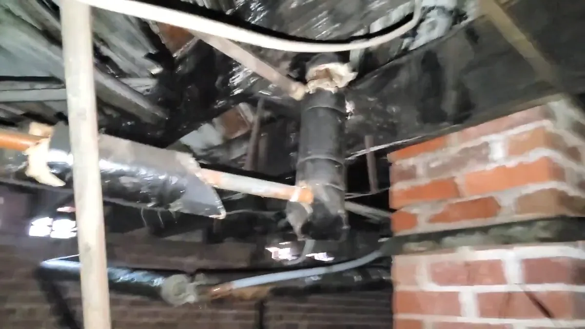 Black wrapped drain and supply pipes running past a red brick pier in a crawlspace