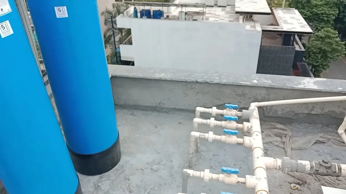 Two tall blue tanks beside a manifold of white pipes and blue-handled valves on a flat roof