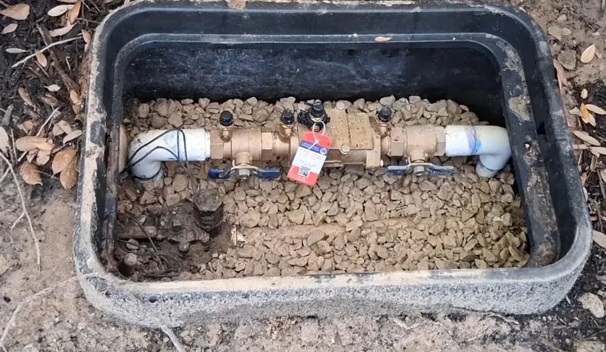 Rectangular ground box holding a brass valve assembly with blue handles, a red tag and a bed of gravel