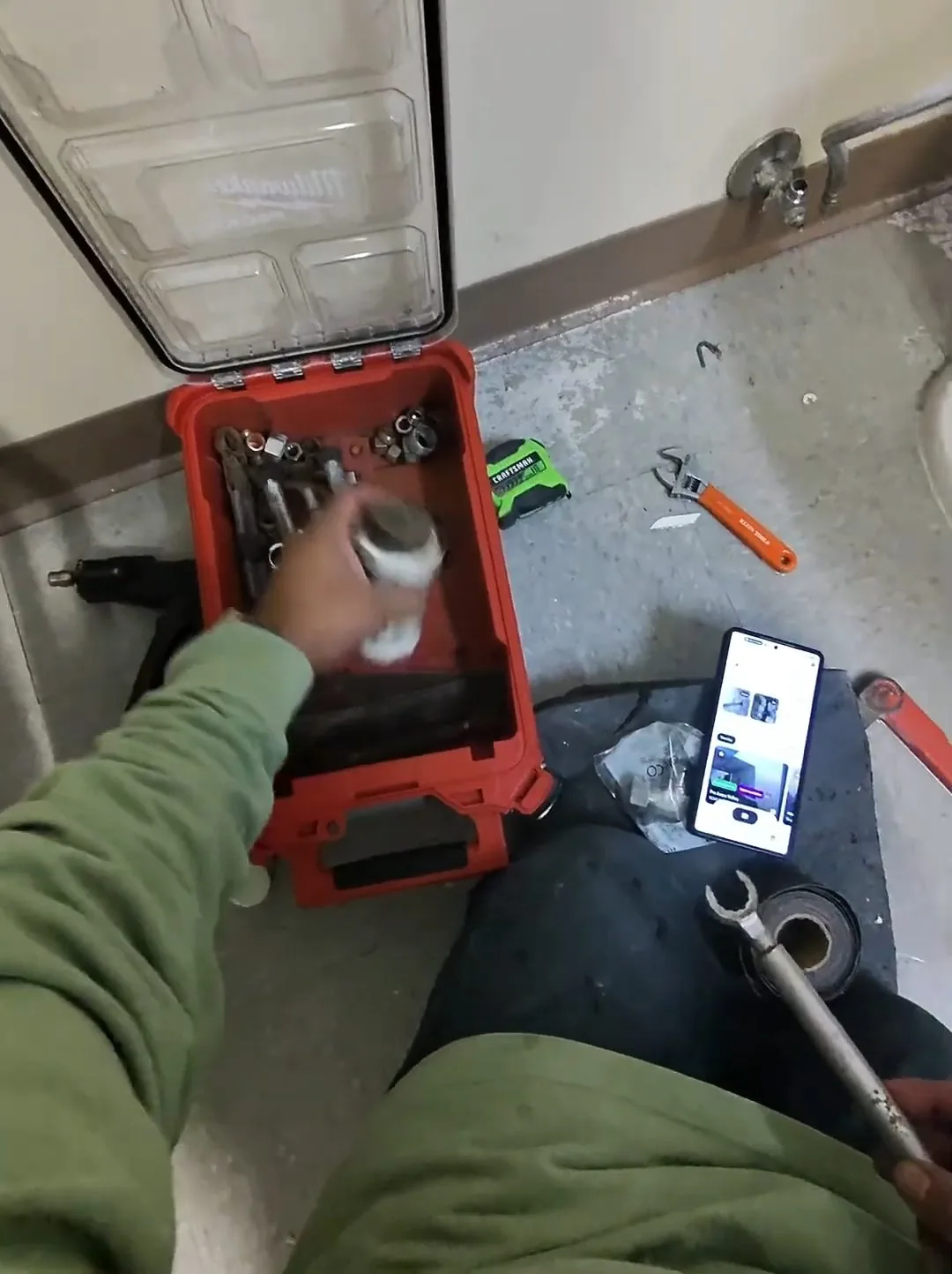 Person kneeling beside a floor drain in a commercial restroom with a red tool case, a wrench and a phone on the floor