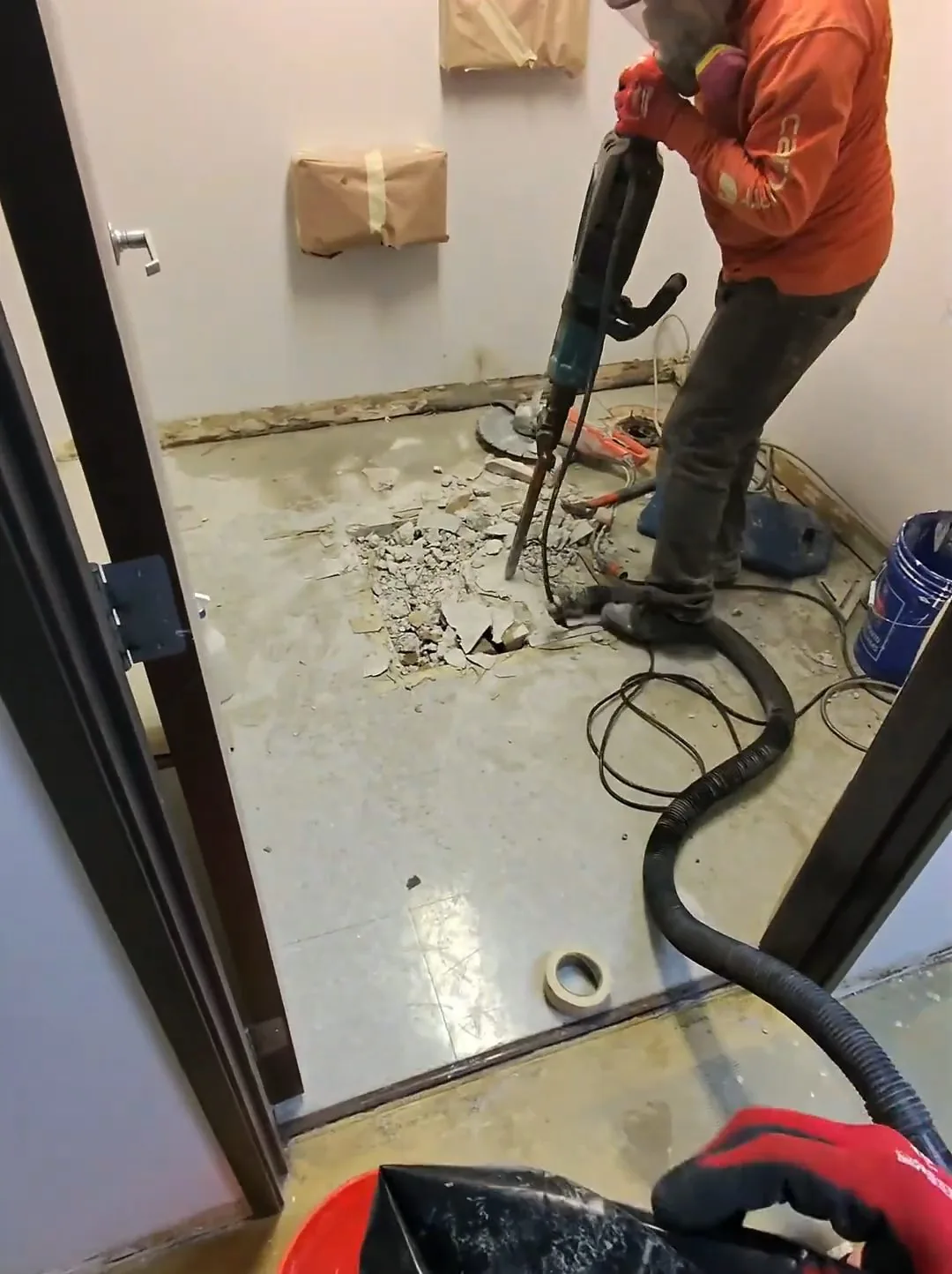 Worker in an orange jacket using a breaker on the floor of a commercial restroom beside a vacuum hose
