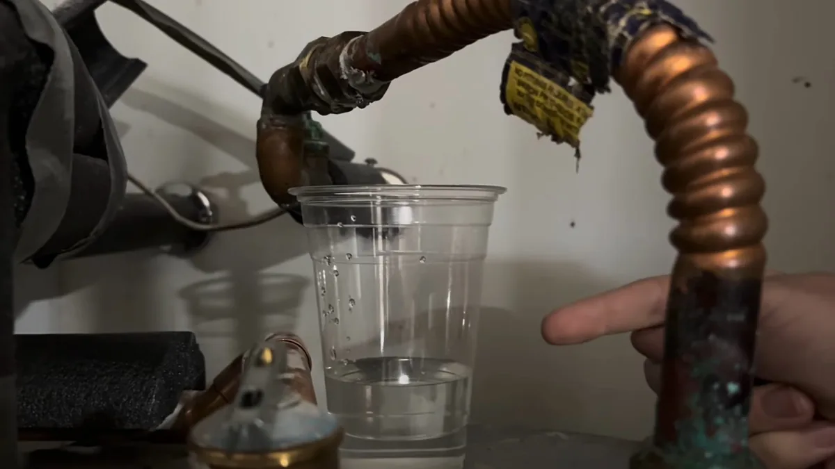 Corroded copper pipe and a corrugated connector at the top of a water heater with a clear cup of water beside them