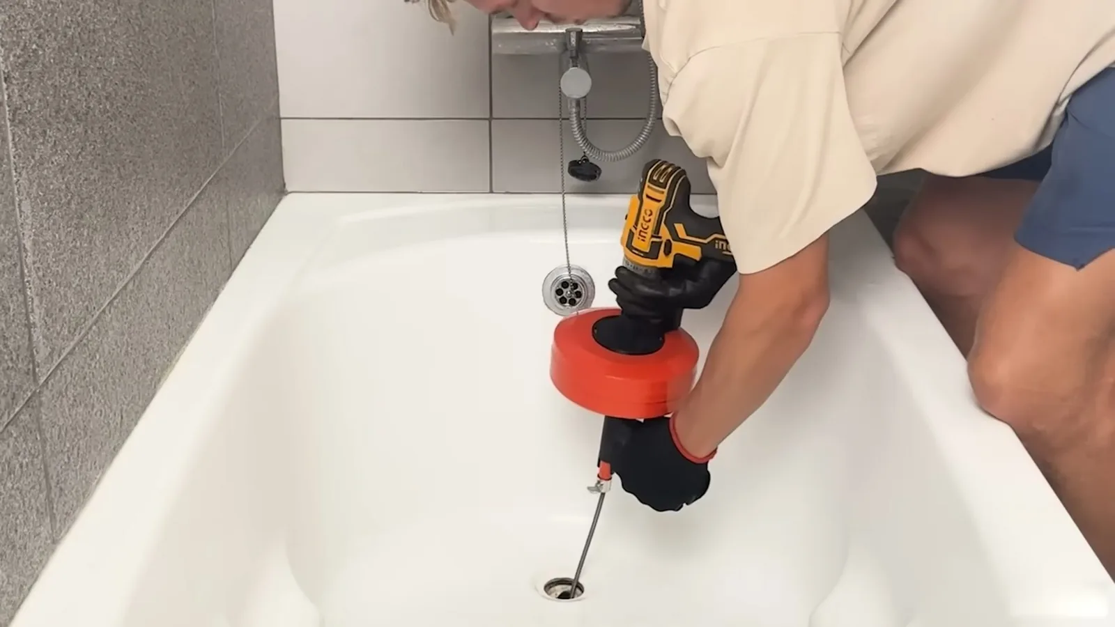 Person leaning into a bathtub to run a drill-driven drain auger with an orange drum into the drain