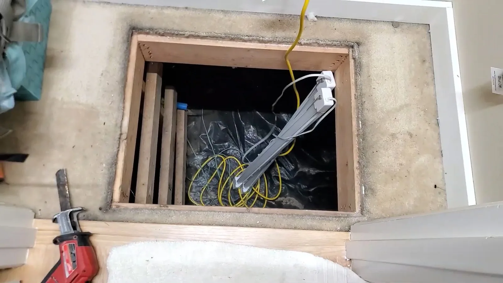 Opened wooden floor hatch over a crawlspace with a work light, a reciprocating saw and a towel beside it