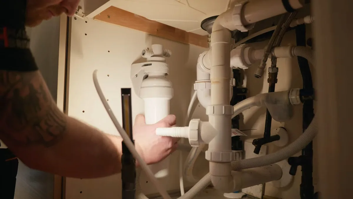 Forearm reaching into a sink cabinet to hold a white drain trap and branch pipe beneath the basin