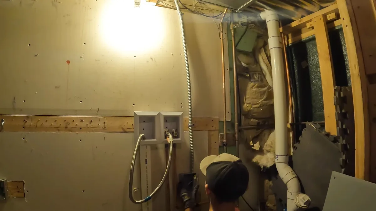 Framed wall with a washing machine outlet box and a white drain pipe, with a worker looking on