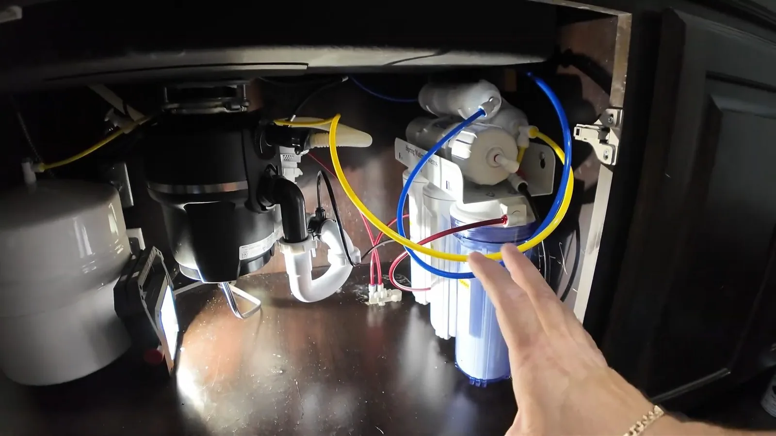 Garbage disposal and water filter hardware mounted in a dark sink cabinet with a hand pointing at the tubing