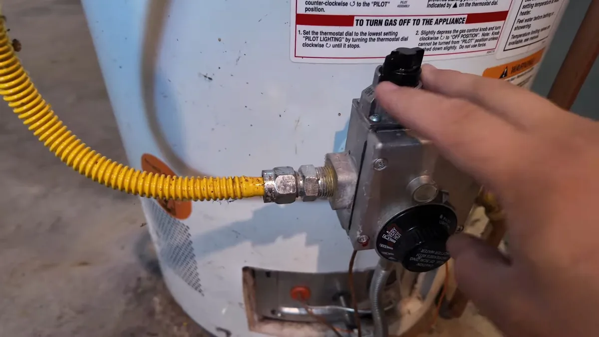 Hand turning the control knob on the gas valve of a tank water heater, with a yellow gas line and warning label beside it