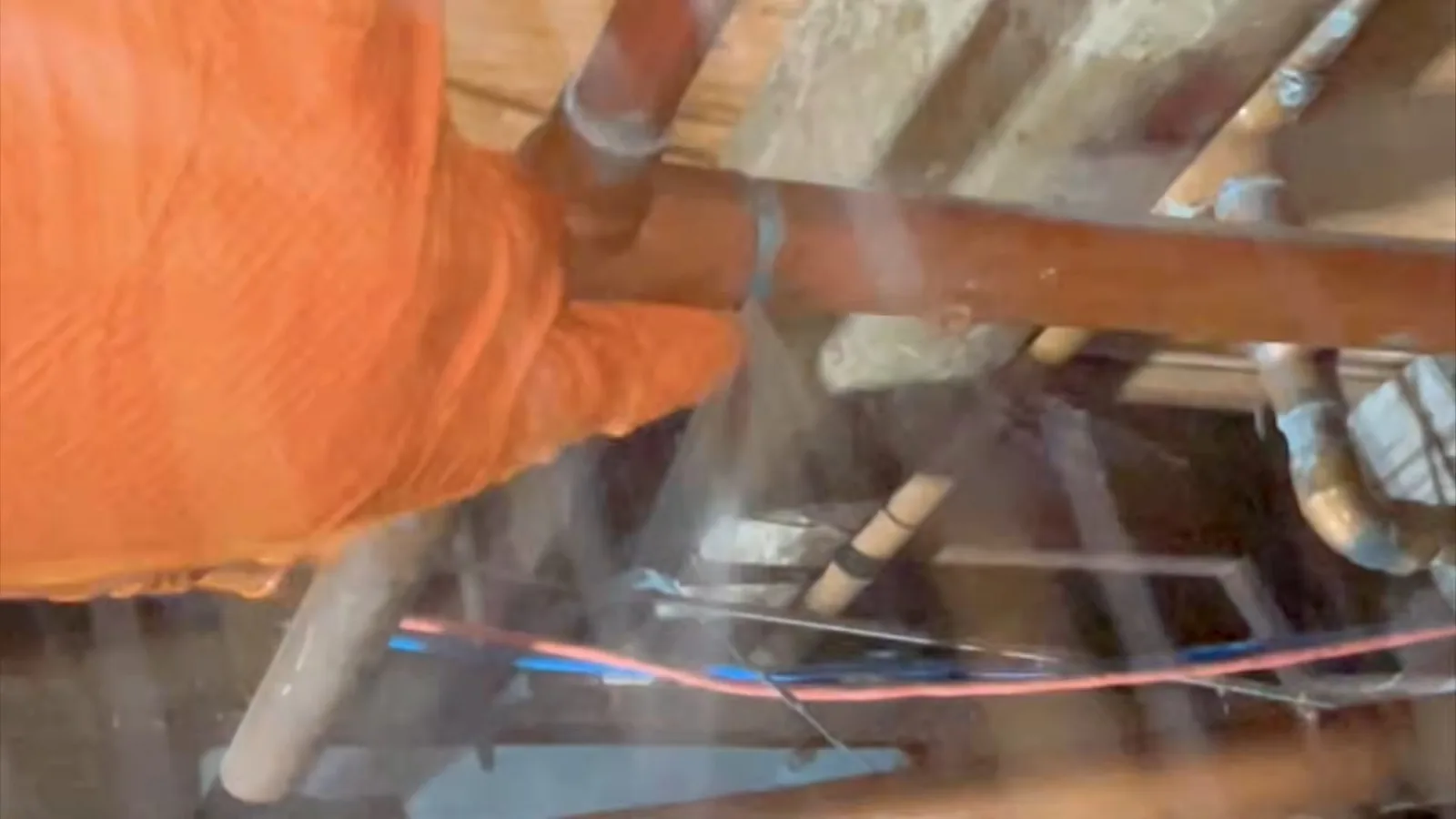 Orange-gloved hand near a pipe in a crawlspace with a fine spray of water coming off the joint