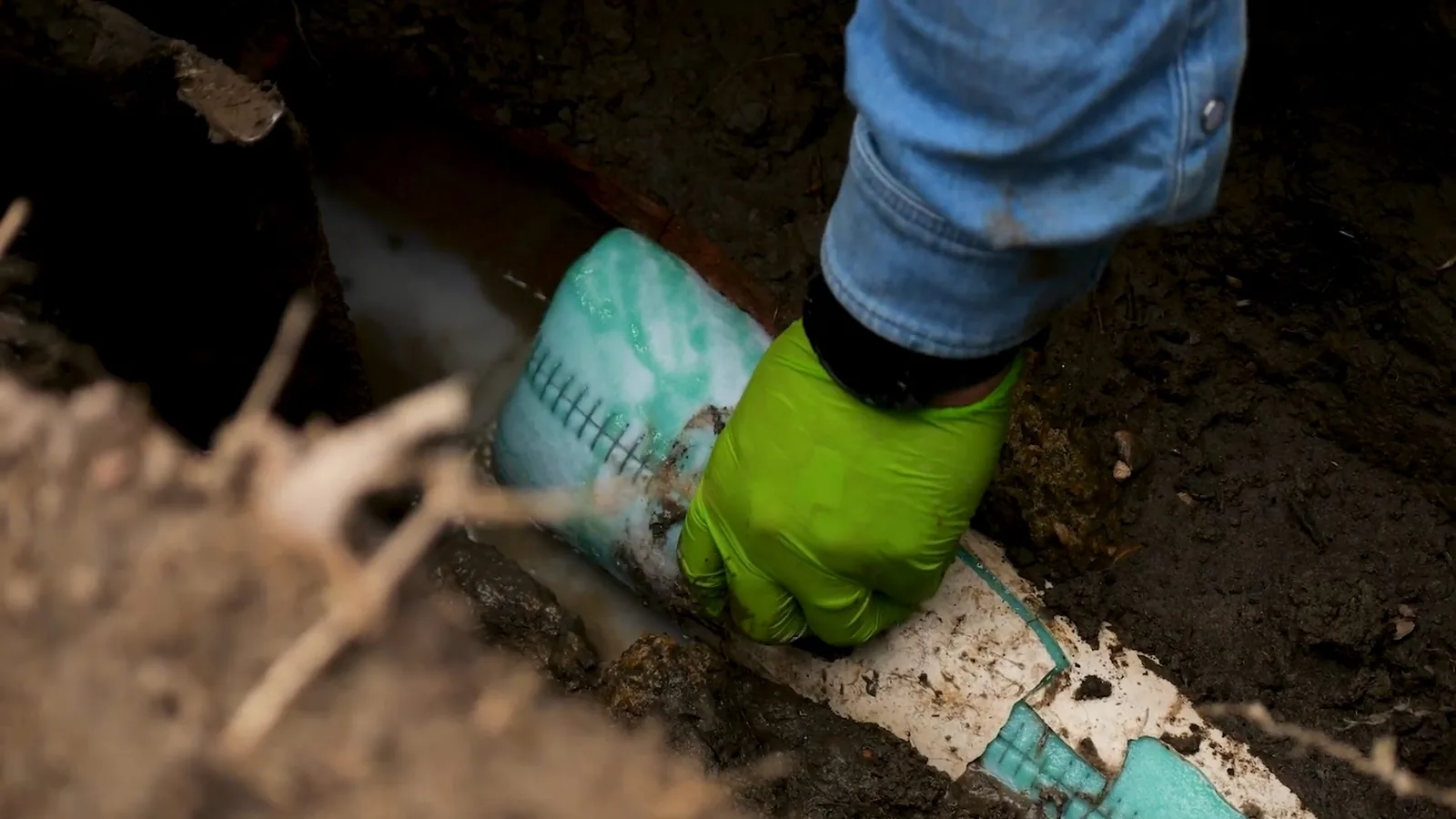 Gloved hand pressing a teal pipe liner into place in an earth excavation