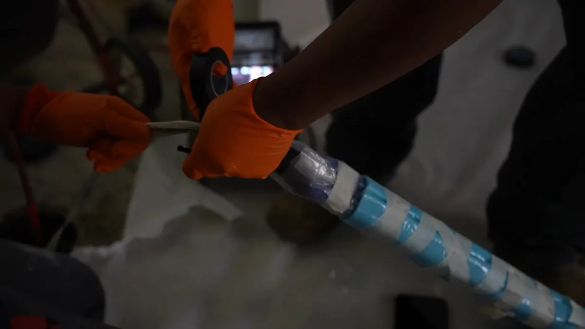 Gloved hands binding a blue-banded liner onto a length of pipe in dim light