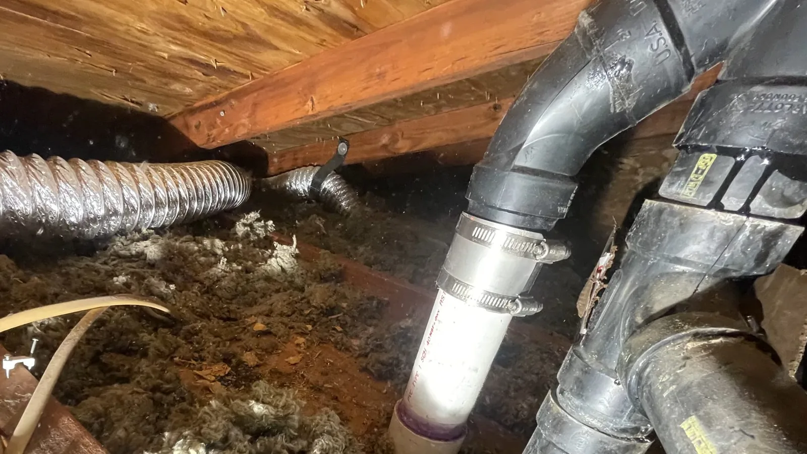 Gray drain and vent pipe with a white clamp coupling running beside a flexible duct under wooden joists