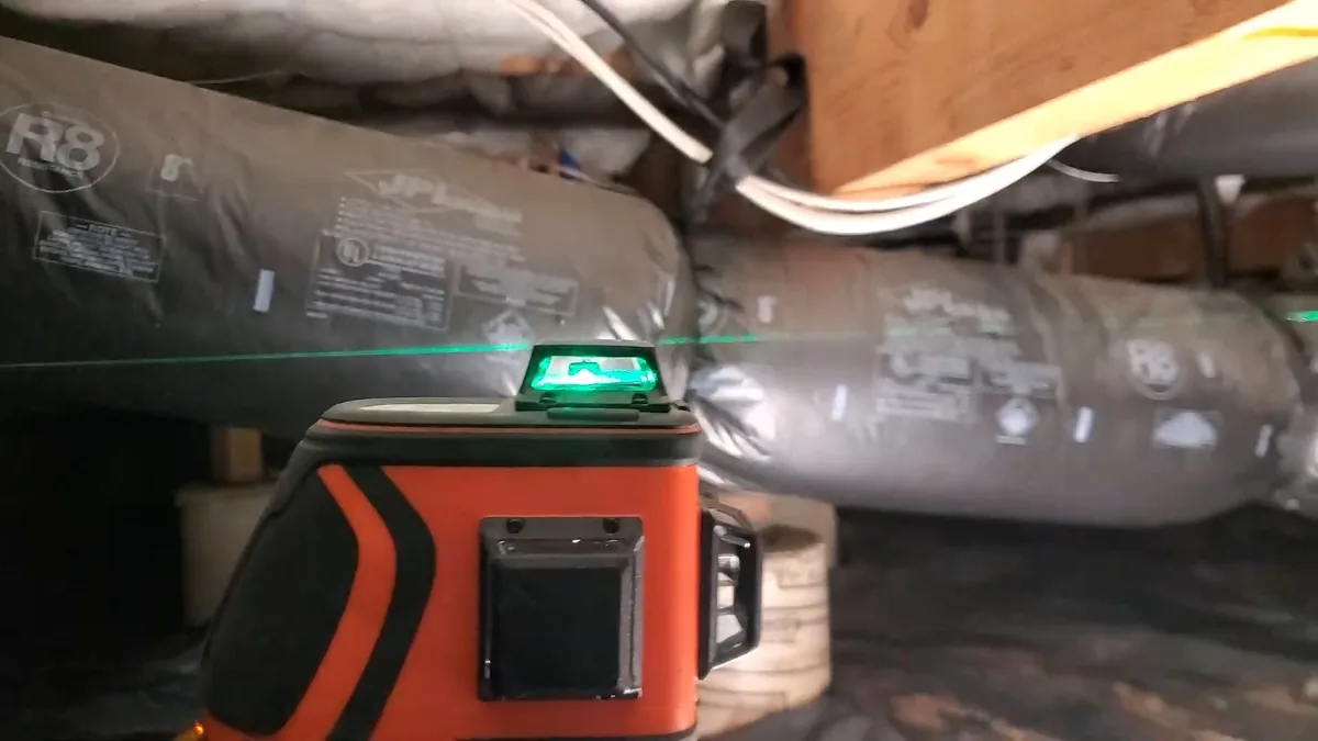 Orange laser level on a crawlspace floor throwing a green line across insulated ducts