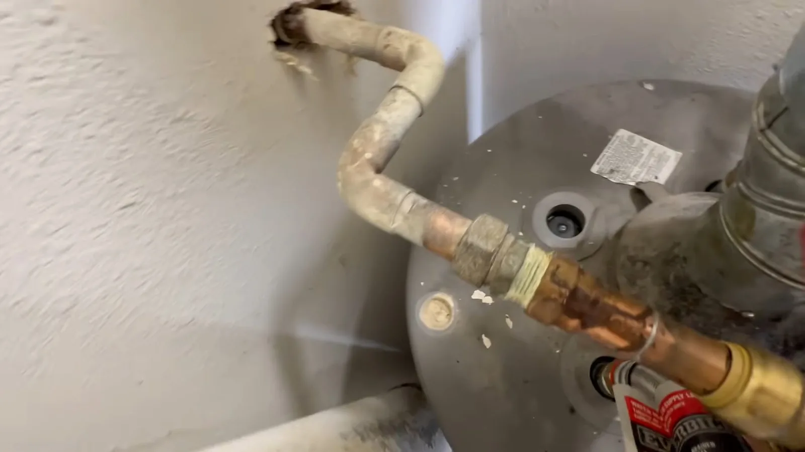 Hand at a corroded flexible connector and copper fitting on top of a gray water heater against a textured wall