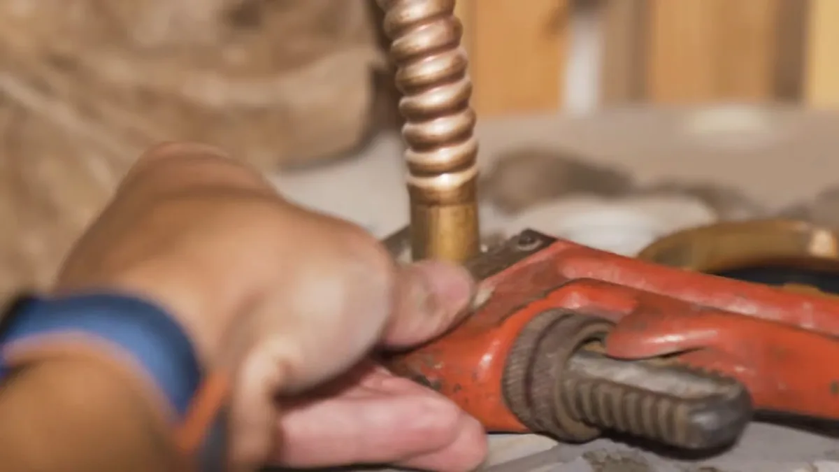 Hand gripping an orange pipe wrench on a brass fitting at the end of a corrugated copper connector