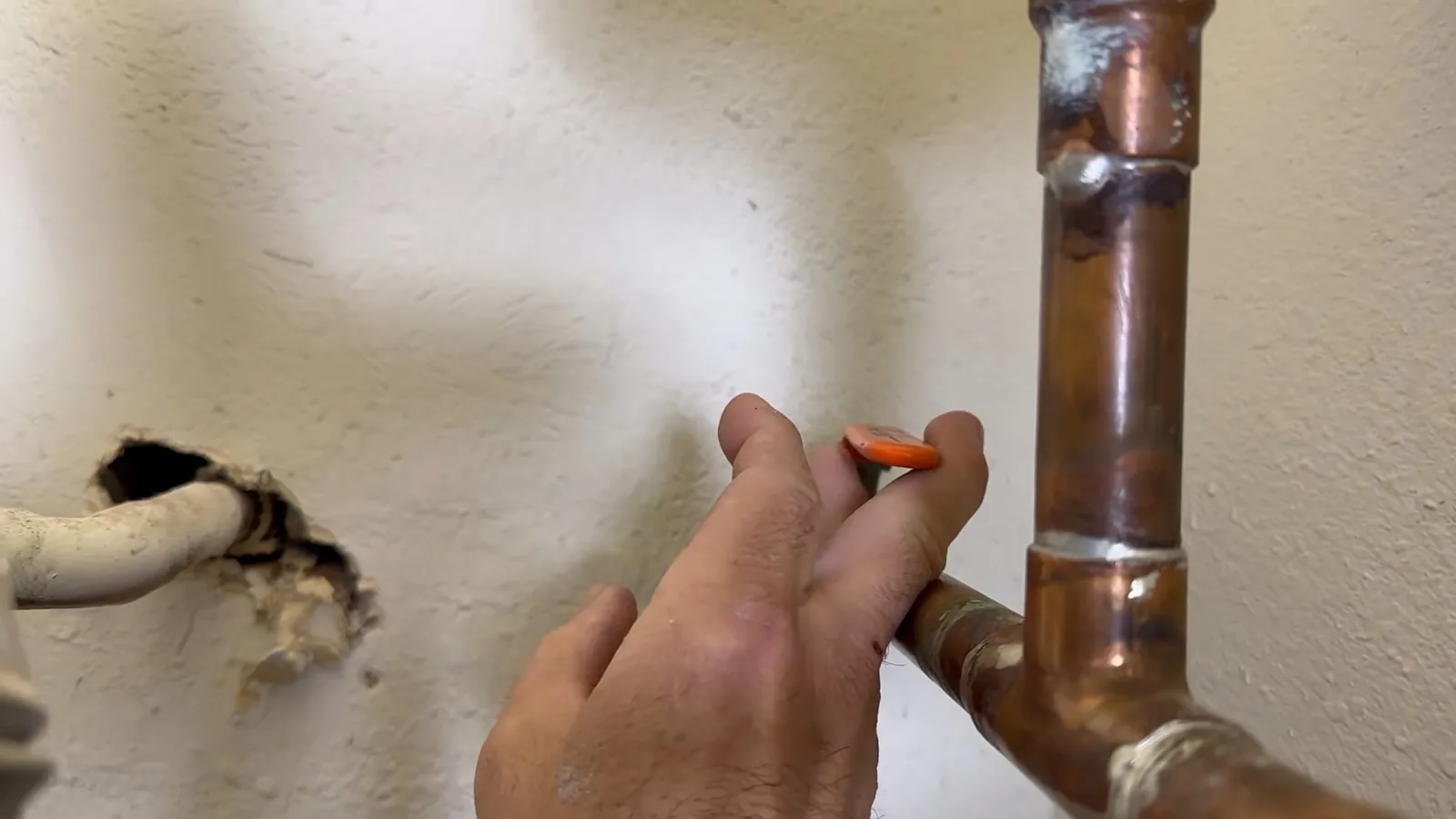 Hand pointing at a copper pipe elbow where the pipe leaves a hole in a painted wall
