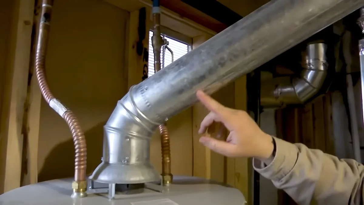 Hand pointing at the angled metal flue on top of a gas water heater with a flexible gas line beside it