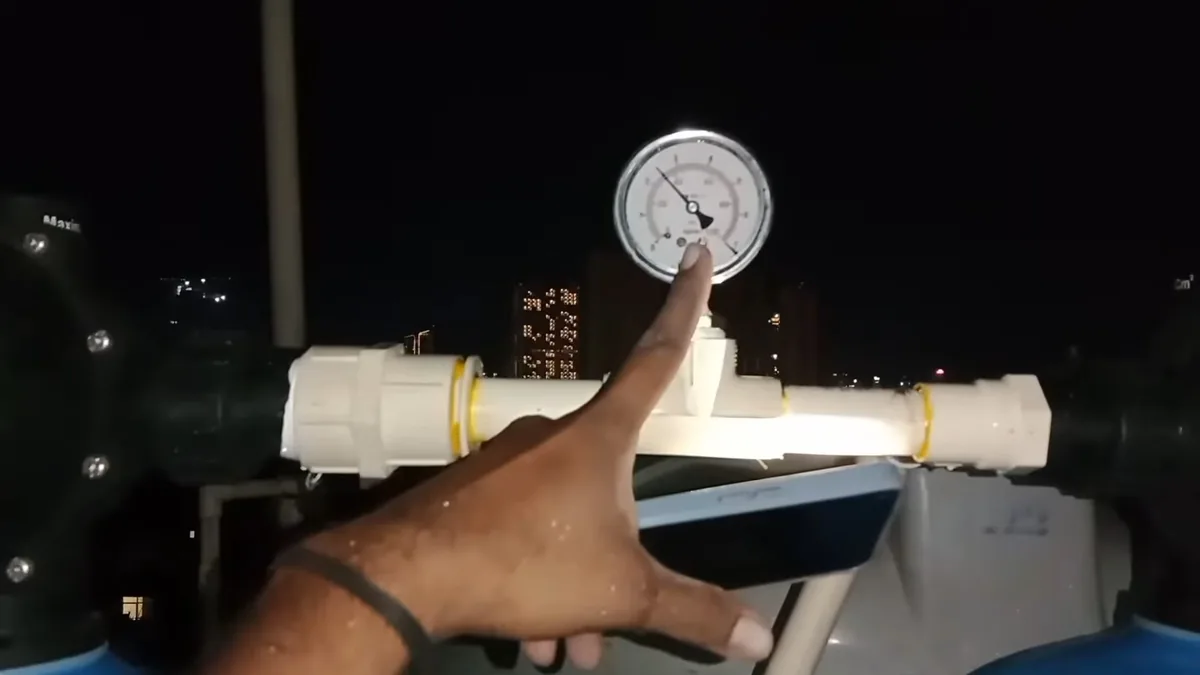 Hand pointing at the dial of a round pressure gauge on a white pipe against a dark background