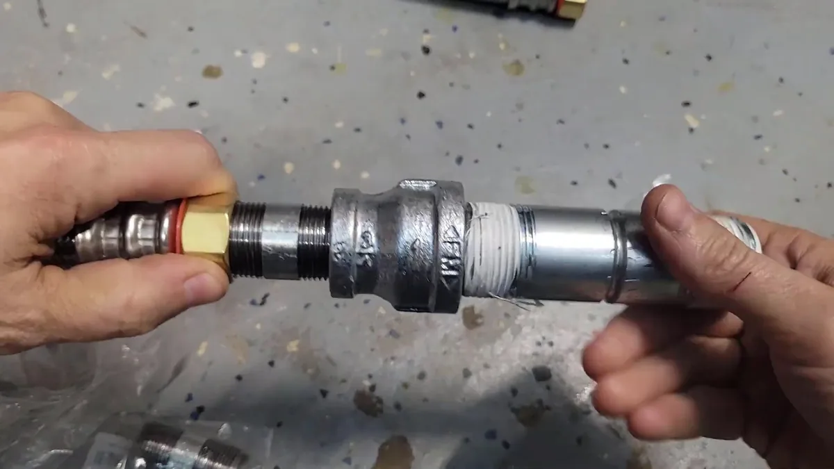 Two hands joining a black iron coupling to a threaded pipe, with a gas connector in the left hand