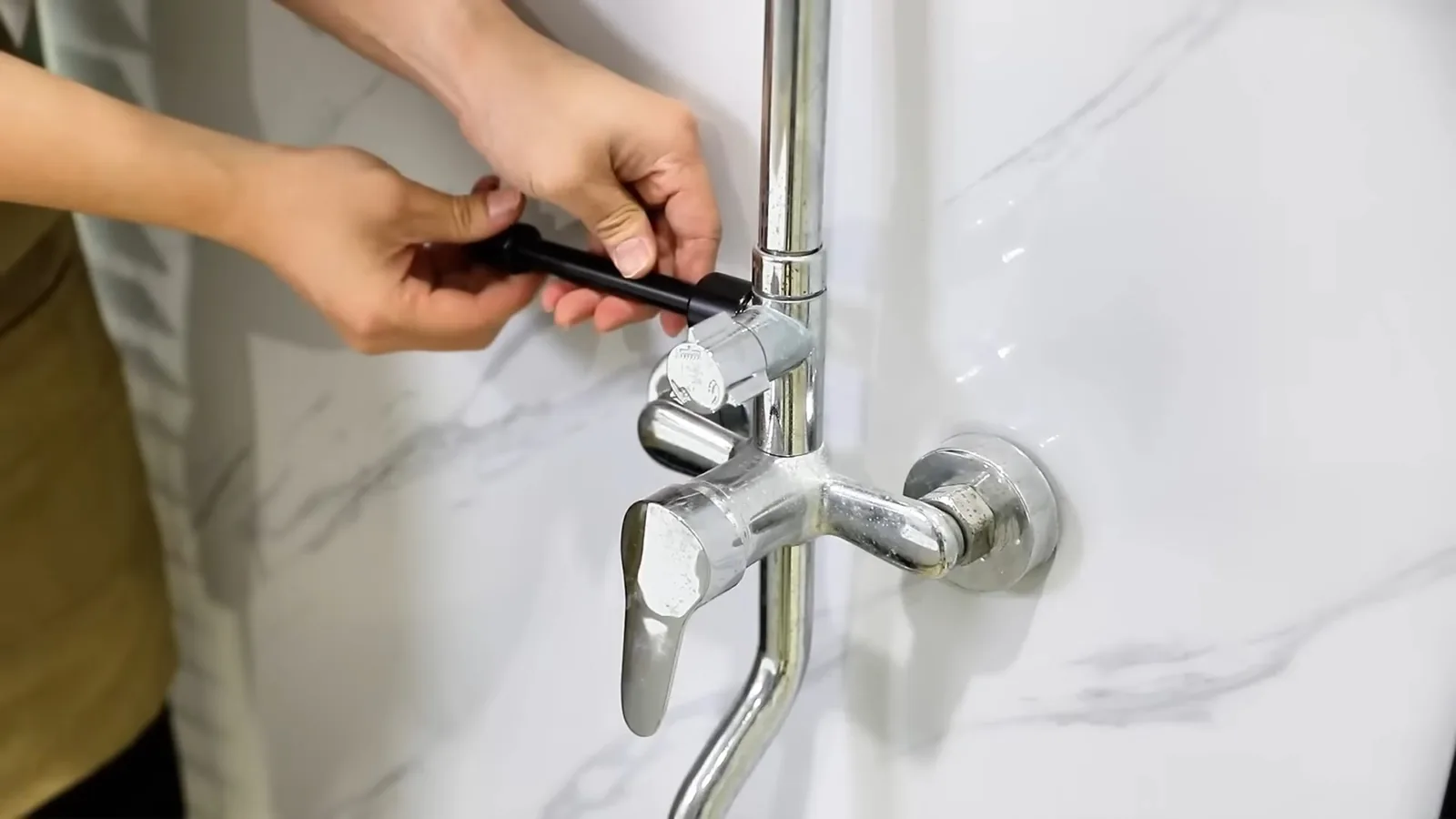 Hands tightening a chrome wall-mounted mixer tap against a white marble-pattern wall
