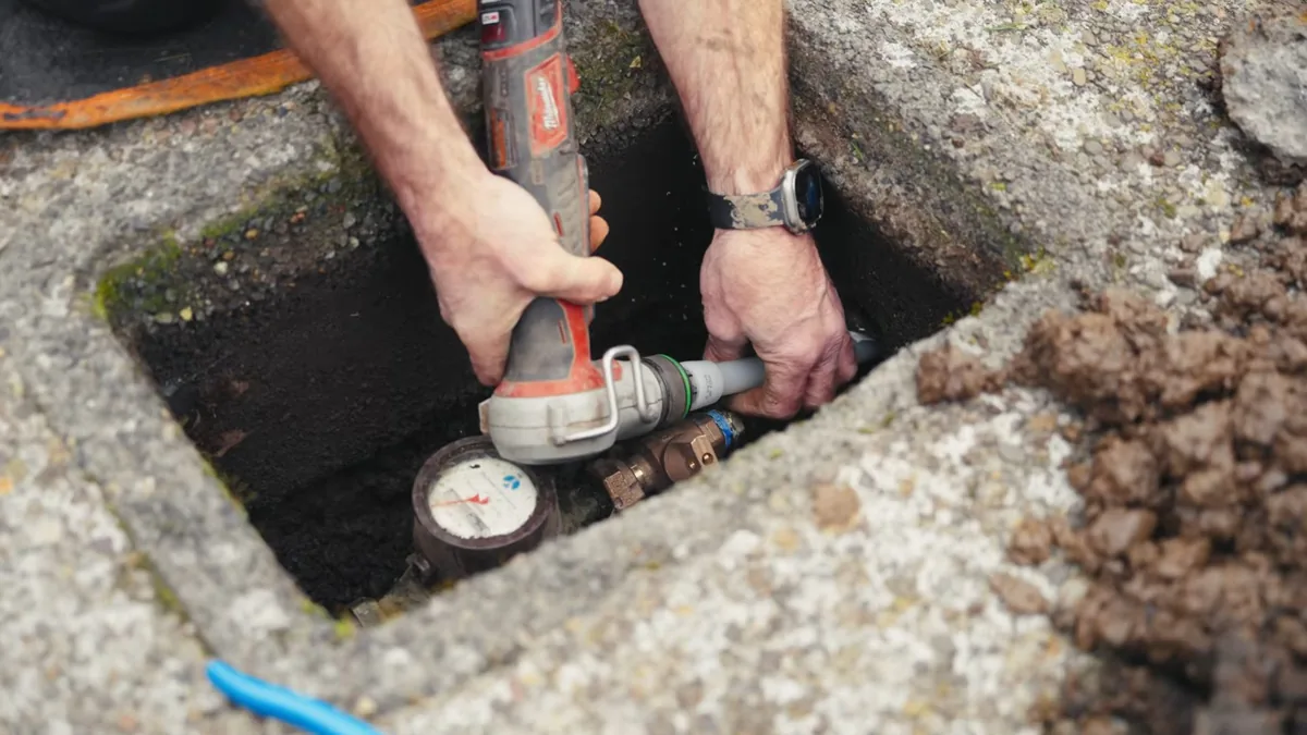 Hands holding a power tool at brass fittings beside a water meter inside a concrete ground box