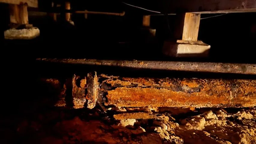 Heavy orange rust and scale along an old cast iron drain pipe beneath wooden floor joists