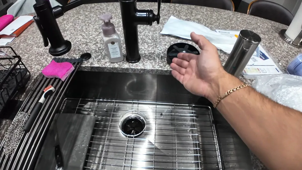 Stainless kitchen sink with a drain strainer, loose fittings laid beside it and a hand reaching in