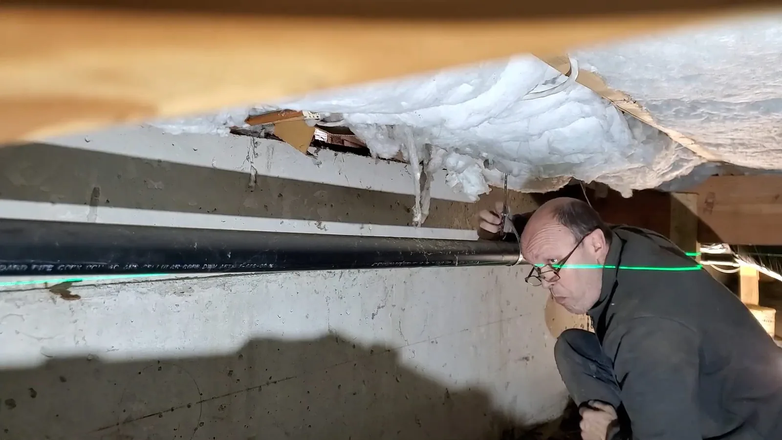 Man crouched at a crawlspace opening beside an insulated ceiling, with a green laser line across a black pipe