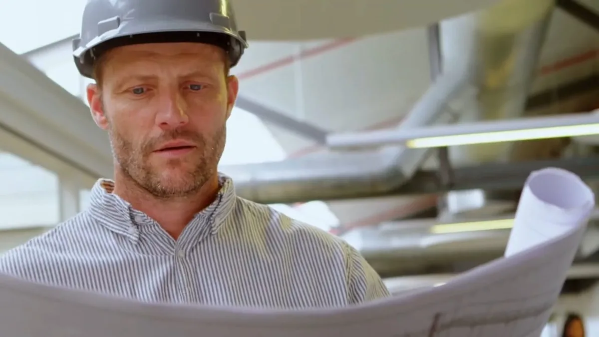 Man in a hard hat reading rolled plans with pipe runs visible behind him