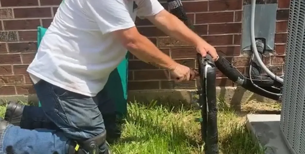 Man kneeling on grass at the side of a brick house, working a black pipe beside an outdoor air conditioner