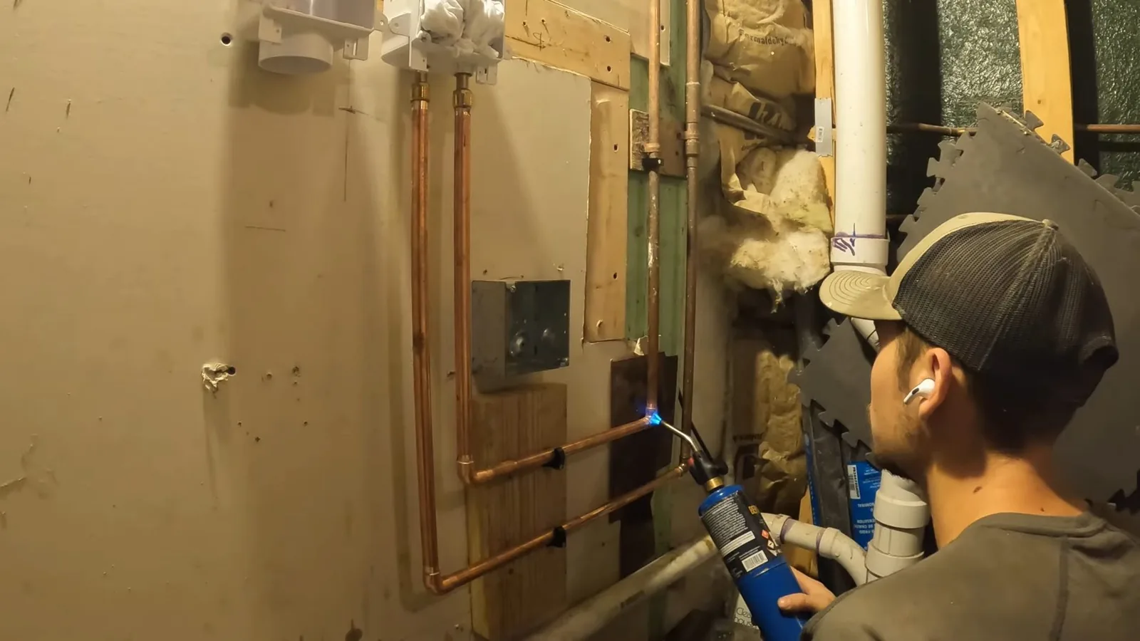 Man in a cap holding a torch to copper lines beside a framed wall with a washing machine box