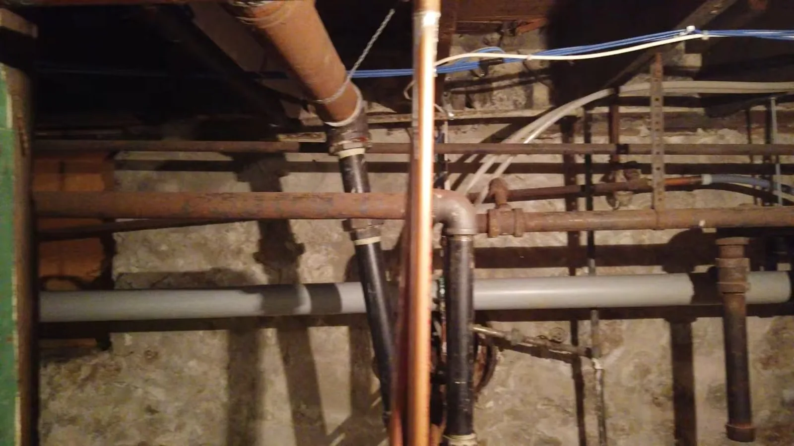 Old copper, iron and plastic pipes crossing beneath exposed joists above a stone foundation wall