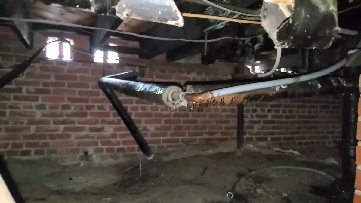 Black and white pipes running along a brick crawlspace wall beneath wooden floor joists