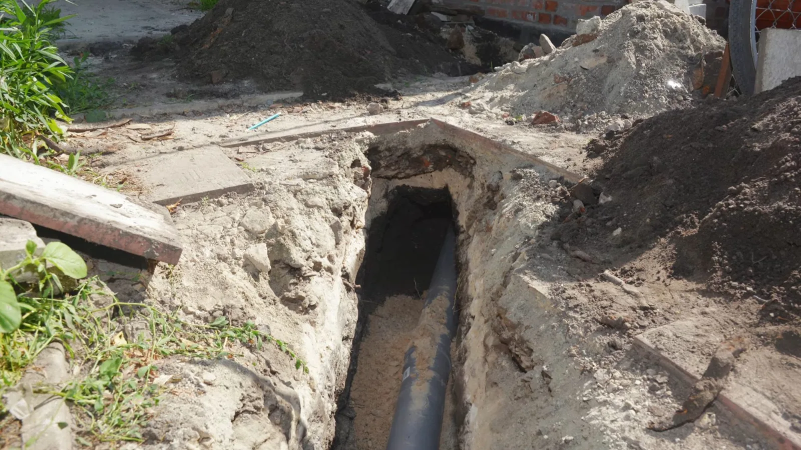 Narrow trench dug beside a house with a gray sewer pipe laid in sand at the bottom
