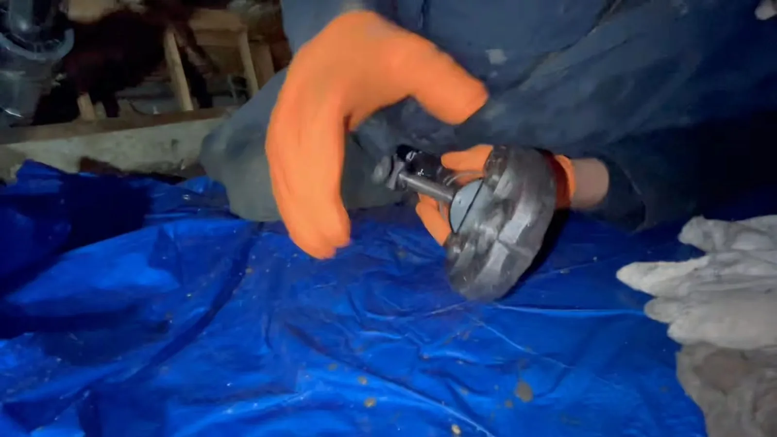 Orange-gloved hands holding a gray flange part over a blue tarp in a crawlspace
