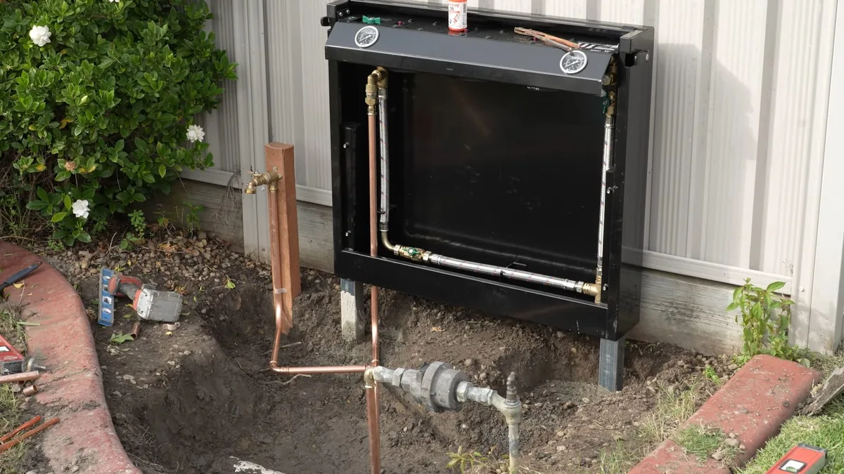Black outdoor equipment box on a siding wall with copper lines and meter fittings at ground level