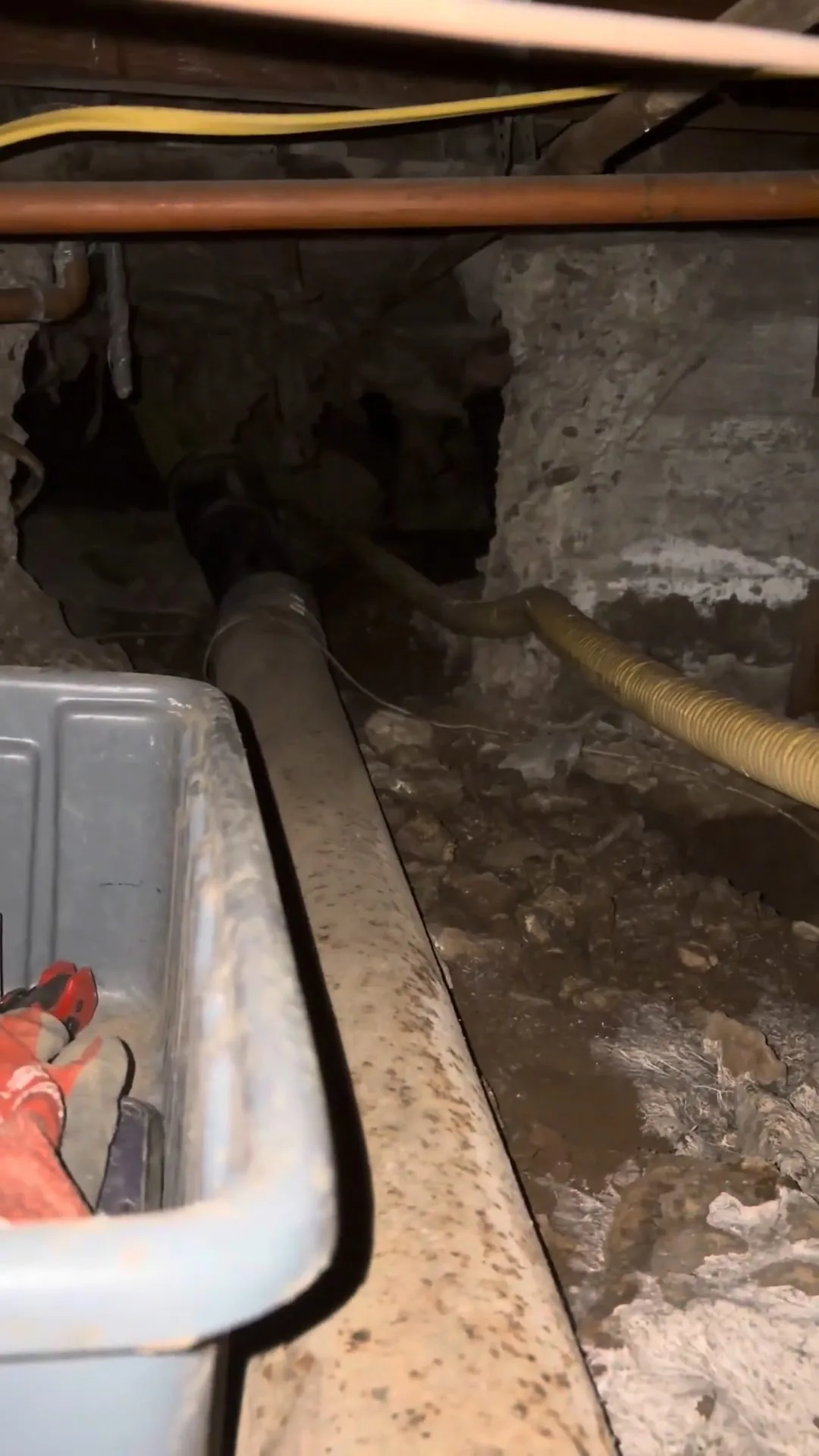 Narrow crawlspace with a pale sewer pipe running along the ground beside a gray tray of tools