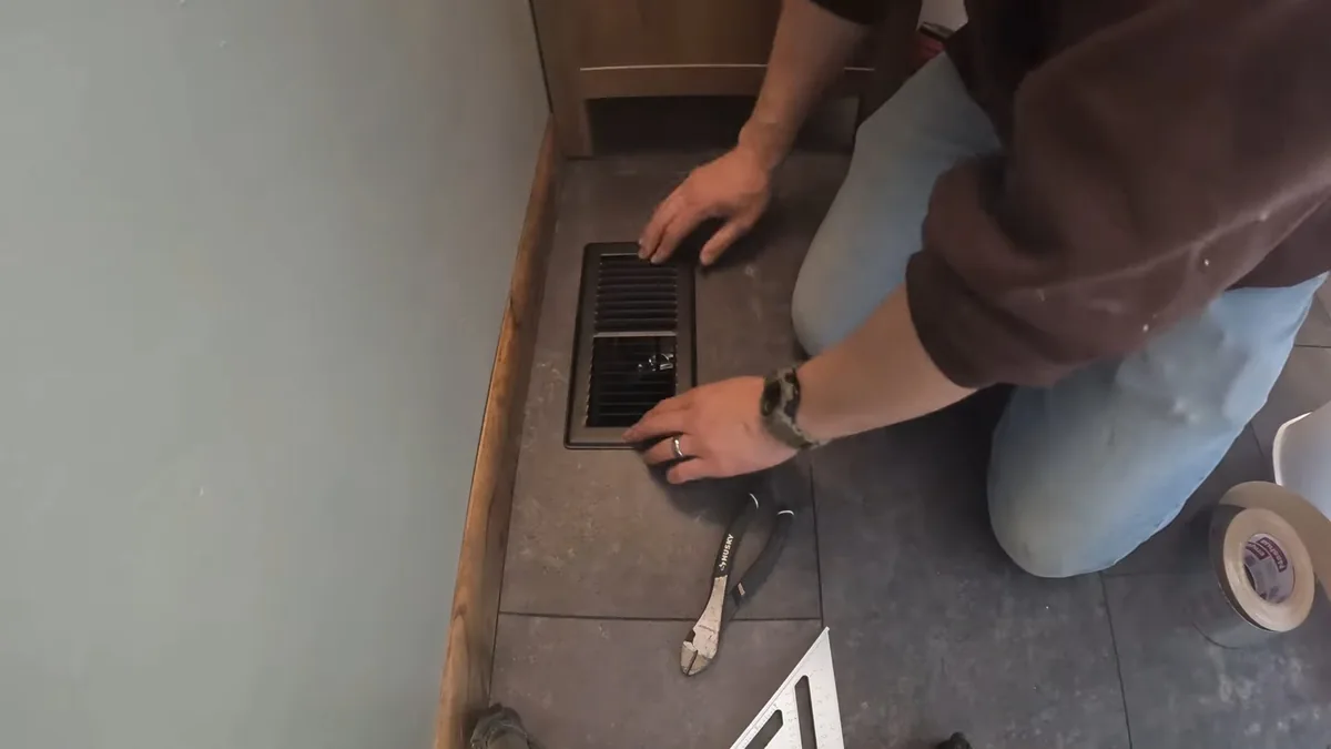 Person kneeling on a tiled floor with a hand on a floor vent grille, pliers and tape beside them