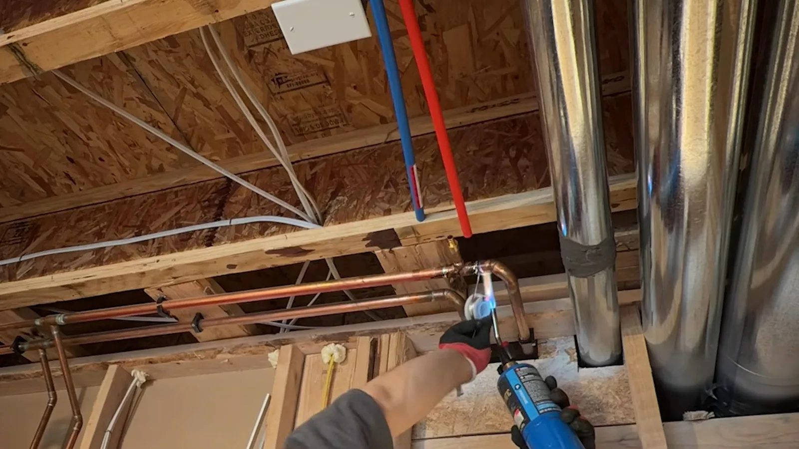 Red and blue PEX supply lines running beside copper pipe being soldered between open floor joists
