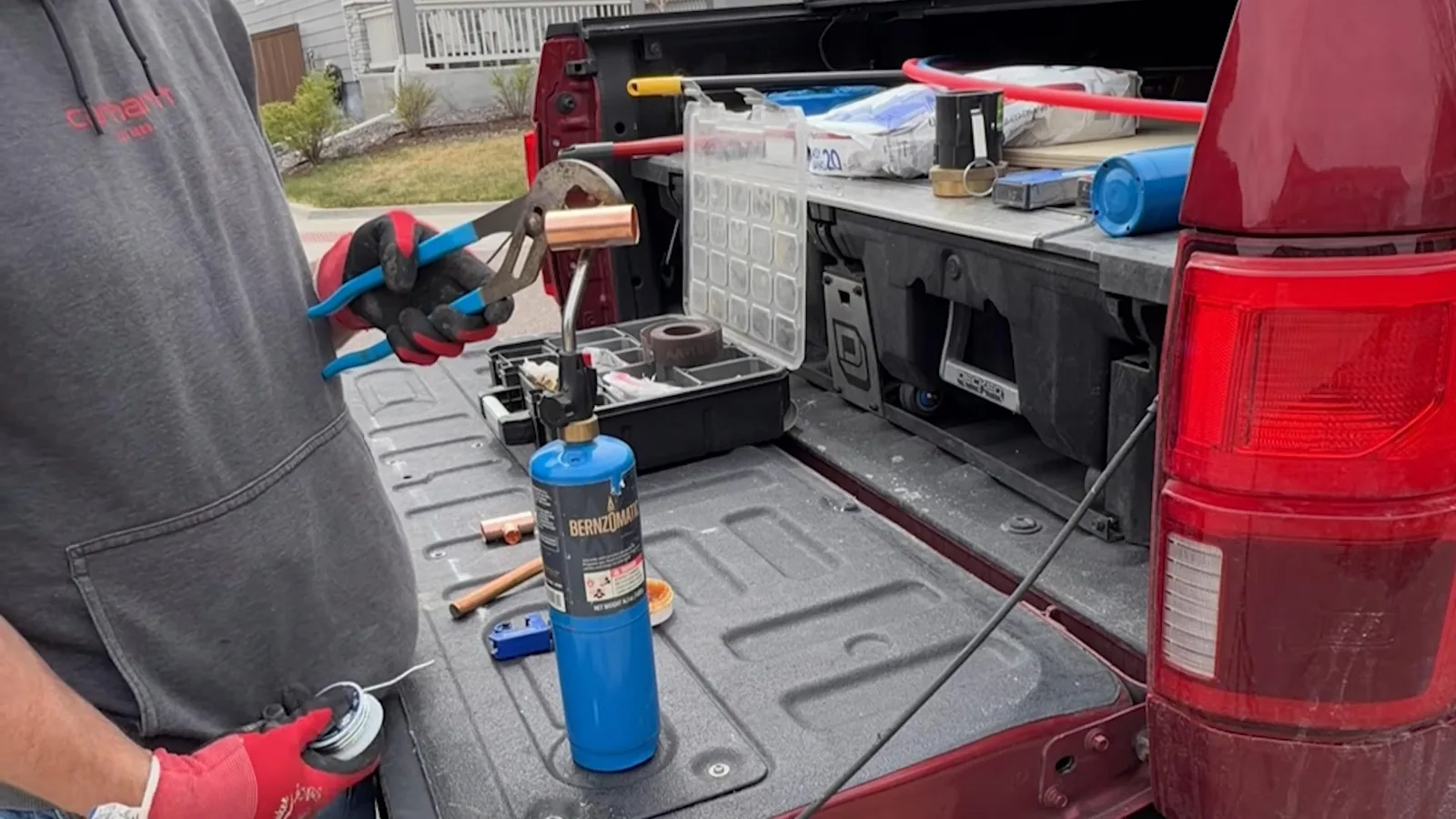 Plumber in a gray sweatshirt holding pliers beside the open bed of a red pickup carrying a torch, a tool tray and fittings