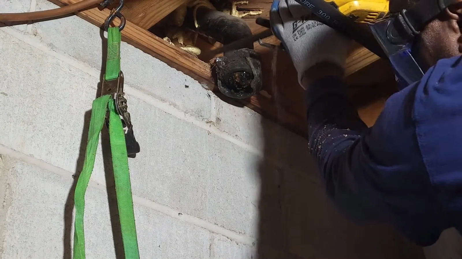 Plumber in blue coveralls using a power saw on a pipe under floor joists beside a block wall