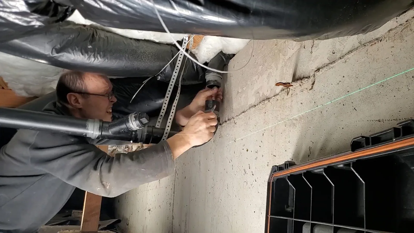 Smiling plumber reaching to fit a pipe hanger on a concrete foundation wall beside a laser line