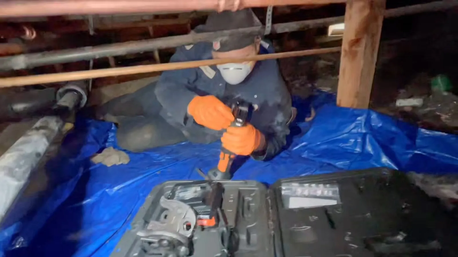 Plumber in a mask and orange gloves working in a crawlspace on a blue tarp beside an open tool case