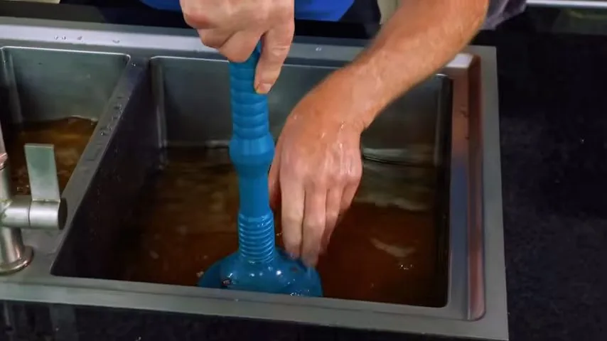Hands working a blue plunger in a stainless sink full of cloudy brown water