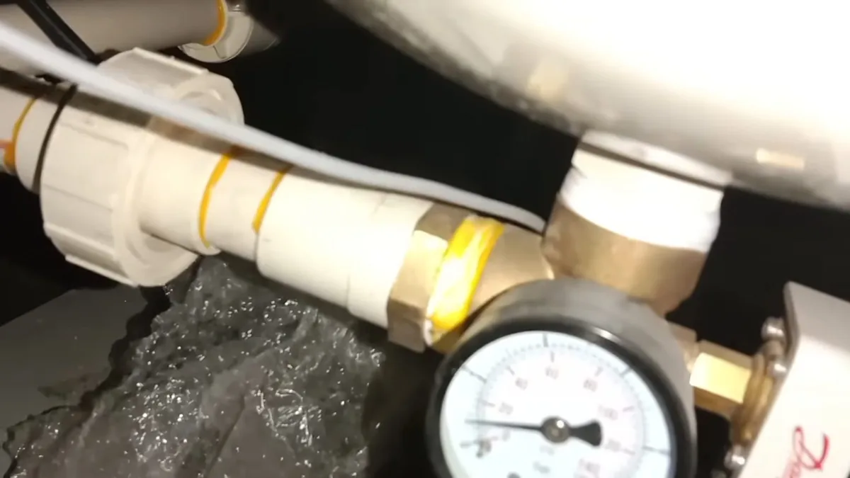 Round pressure gauge on a brass fitting at a white pipe, with water running underneath