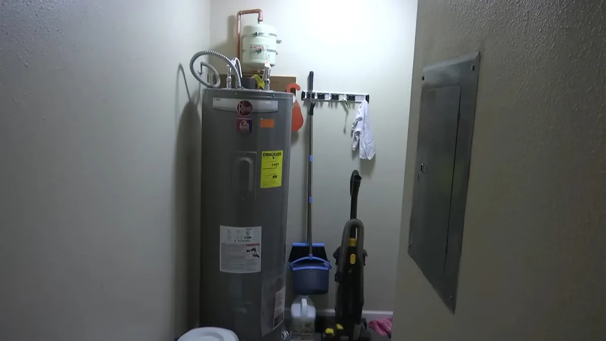 Gray electric tank water heater standing against the wall of a utility room with a supply pipe running overhead