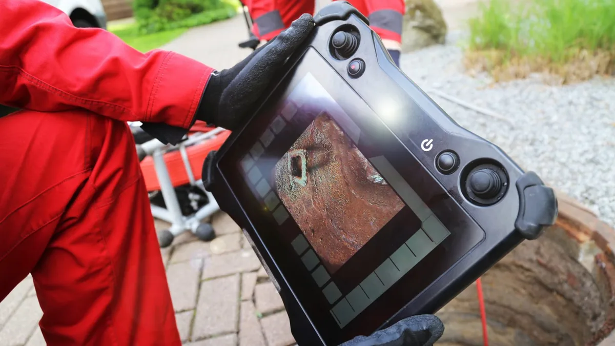 Technician in red coveralls holding a handheld camera monitor with a pipe wall on screen, a manhole and cable reel behind
