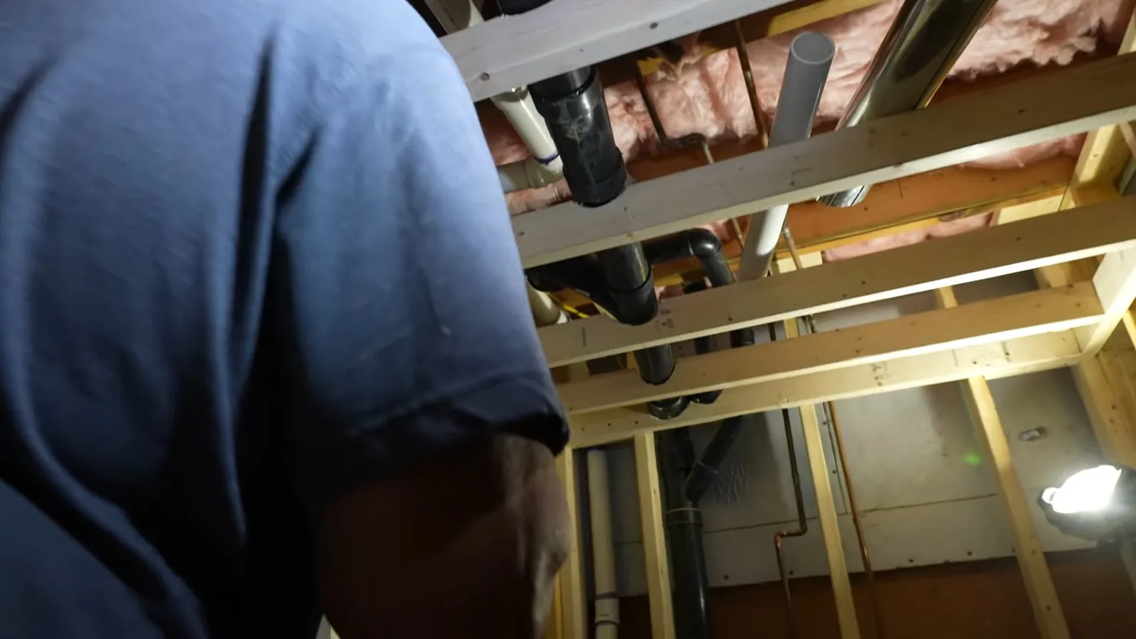 Technician in a blue shirt looking up at drain and vent pipes through open basement framing